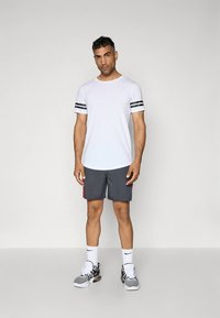 Camiseta branca de manga curta com riscas pretas nas mangas, calções cinzentos com detalhes vermelhos e ténis desportivos cinzentos com meias brancas.