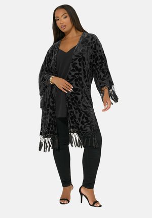 Black velvet kimono with leaf pattern, fringed edges, open front, worn over a black top and pants, featuring high-heeled black sandals.