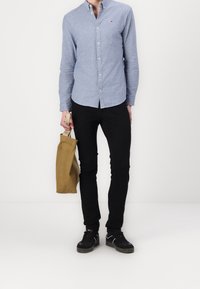 Jasnoniebieska koszula z długim rękawem z guzikami, czarne spodnie slim-fit, czarne trampki, trzymając brązową torbę z tkaniny. Minimalistyczny design, casualowy styl.
