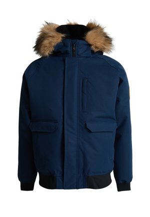 Dreng med flettet hår iført en mørkeblå vinterjakke med pelsforet hætte, grå joggingbukser og hvide sneakers står foran en ensfarvet baggrund.