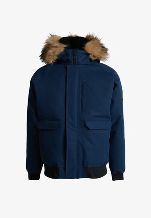 Jongen met gevlochten haar, gekleed in een marineblauwe winterjas met bontkap, grijze joggingbroek en witte sneakers, staand tegen een effen achtergrond.