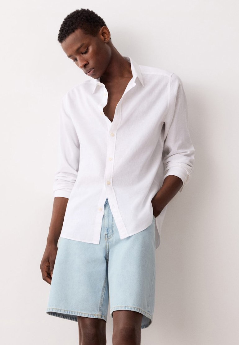 Homme portant une chemise blanche à manches longues boutonnée et un short en jean bleu clair, debout avec une main dans la poche devant un fond blanc.