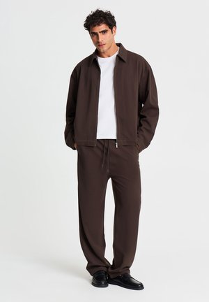 Uomo con capelli ricci che indossa giacca e pantaloni marrone scuro, maglietta bianca e scarpe nere, in piedi davanti a uno sfondo semplice.