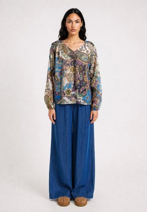 Woman wearing a long-sleeve floral patterned blouse, wide-leg blue denim pants, and brown flat shoes, standing against a plain background.