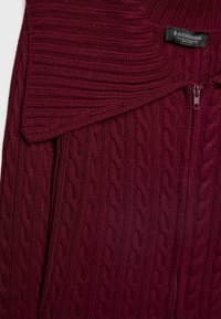 Bordeauxrode gebreide cardigan met kabelgebreide textuur, voorzien van een omgeslagen kraag en een ritssluiting. Stradivarius merklabel zichtbaar.