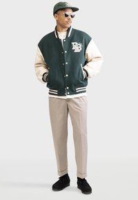 Grüne und cremefarbene Collegejacke mit Logo, schwarzen gestreiften Bündchen, beigen Hosen und schwarzen Schuhen. Die Accessoires umfassen Sonnenbrillen und eine passende Mütze.