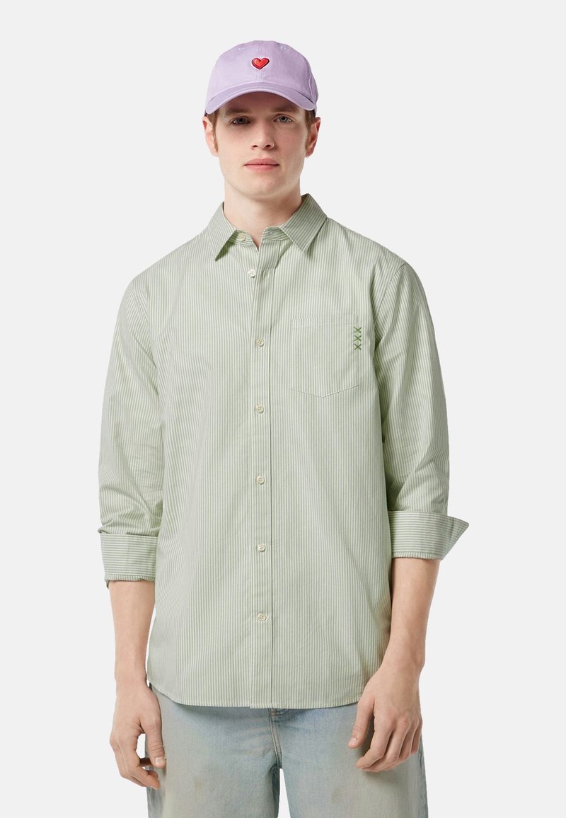 Camicia a righe verde chiaro con taschino sul petto e dettagli ricamati, abbinata a un cappellino lavanda con un design a forma di cuore rosso.