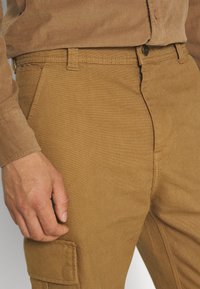 Primer plano de una persona con camisa marrón de pana y pantalones cargo color marrón claro con bolsillo lateral y presillas visibles.