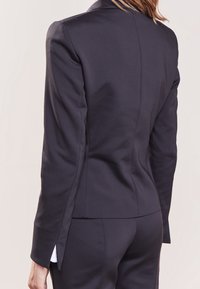 Blazer noir sur mesure en tissu lisse, présentant une silhouette ajustée, des épaules structurées et des petites fentes latérales à l'ourlet.