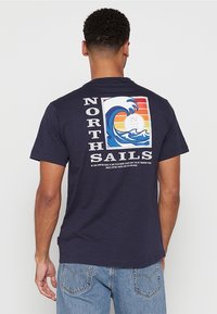 Ciemnoniebieski bawełniany t-shirt z dużą grafiką przedstawiającą fale i zachód słońca oraz napisem "NORTH SAILS" w białym kolorze.