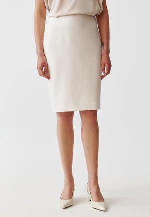 Femme portant une jupe crème ajustée au niveau des genoux et des chaussures à talons hauts crème assorties, debout devant un fond blanc uni.