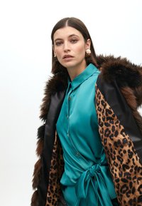 Tirkizna satenska bluzica s detaljem gumba i kravatom u struku, uparena s kaputom od umjetnog krzna u leopard uzorku crno-smeđe boje.