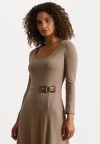 Donna con capelli ricci che indossa un vestito beige a maniche lunghe con un dettaglio di cintura a doppio fermaglio marrone in vita, su uno sfondo neutro.