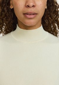 Svetlo bež pulover z visokim ovratnikom in rebrastim tkaniem. Poudarjen bližnji posnetek, ki prikazuje obliko ovratnika in ten kože modela.