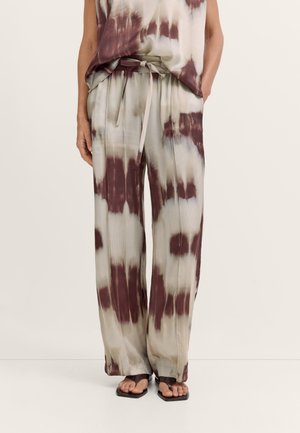 Lockere, weit geschnittene Batik-Hose in Beige und Dunkelbraun mit Kordelzug in der Taille, kombiniert mit schwarzen Sandalen mit offenen Zehen und passendem Oberteil.
