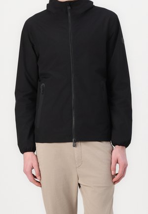 Personne portant une veste noire à capuche zippée avec des poignets élastiques et un pantalon beige, debout devant un fond uni.