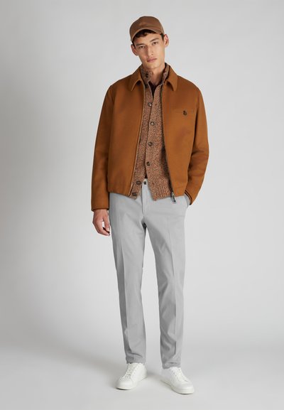 Młody mężczyzna w brązowej czapce, brązowej kurtce na sweterze, jasnoszarych spodniach i białych sneakersach, stojący na jednolitym tle.