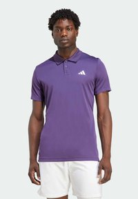 Lila Sport-Polo-Shirt mit kurzen Ärmeln, drei Knöpfen und einem weißen Logo auf der linken Brust. Aus glattem, leichtem Stoff gefertigt.