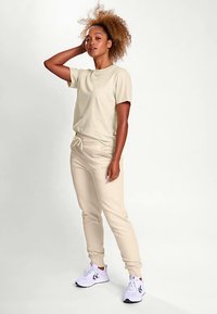 Beiges Baumwoll-T-Shirt mit rundem Ausschnitt, kombiniert mit passenden Jogginghosen mit elastischem Bund und Seitentaschen. Weiße Turnschuhe.