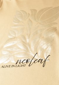 Design de feuille brillante en beige clair avec le texte « neo leaf » en cursive et « ALIVE IN LIGHT » en majuscules sur fond en tissu.