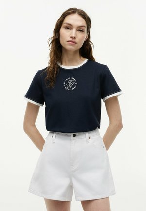 Kvinde med langt brunt hår iført en navyblå Tommy Hilfiger t-shirt og hvide shorts med høj talje, stående foran en ensfarvet hvid baggrund.