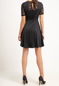 Czarna sukienka w kształcie litery A z krótkimi koronkowymi rękawami, teksturowanym korpusem i detalem w kształcie dziurki na plecach. Gładki materiał i rozkloszowany dół. Wysokie obcasy.