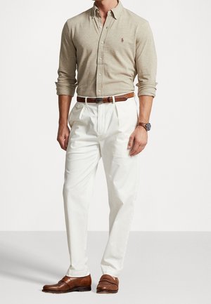 Mand iført beige skjorte med knapper, hvide plisserede bukser, brunt bælte, brune pæne sko og armbåndsur, stående foran ensfarvet baggrund.