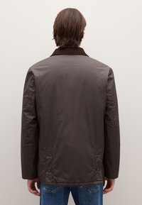 Giacca marrone leggera con colletto a costine, texture liscia e dettagli di cuciture visibili. Presenta un taglio dritto e tasche posteriori.