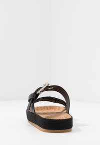 Black sandal with cork footbed, braided black strap, and silver hardware detail. Features a flat sole with a textured finish.