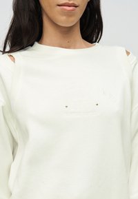 Γυναίκα που φοράει λευκό φούτερ με ανοίγματα στους ώμους και ανάγλυφο κείμενο που αναγράφει «ELISABETTA FRANCHI SINCE 1998 LIMITED EDITION».