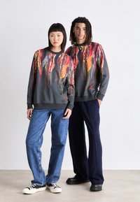 Two individuals wear dark gray sweatshirts featuring vibrant red, yellow, and white abstract designs. They pair with blue jeans and black shoes.