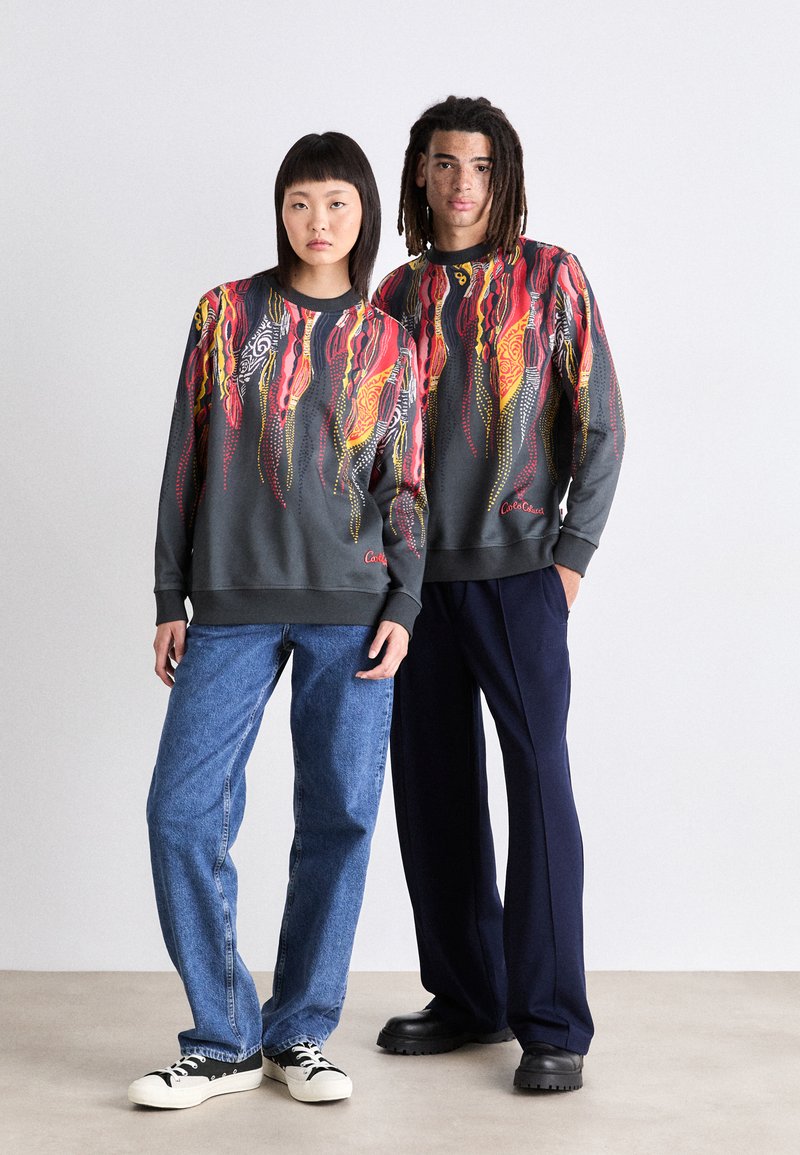 Twee individuen dragen donkergrijze sweatshirts met levendige abstracte ontwerpen in rood, geel en wit. Ze dragen daarbij blauwe jeans en zwarte schoenen.