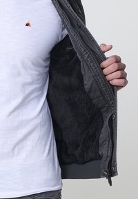 Grå ytterjacka med mjuk svart pälsfoder. Bärs över en vit T-shirt, vilket visar en enkel, stilren design med dragkedja.