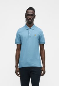 Polo azul claro em algodão, com colarinho, três botões e um pequeno logótipo amarelo no peito esquerdo. Mangas curtas.