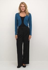 Niebieski krótki kardigan z okrągłymi krawędziami, zestawiony z czarnymi spodniami o wysokim stanie. Gładka faktura, jednolity kolor i luźny krój. Czarne płaskie buty.