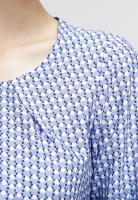Blusa azul clara con escote redondeado, que presenta un patrón de puntos oscuros y blancos. Tela ligera con textura suave.