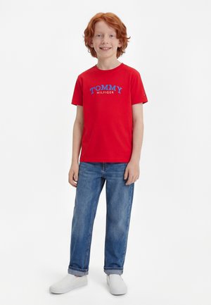 Dječak s crvenom kosom smiješi se, nosi crvenu Tommy Hilfiger majicu, plave traperice s manžetama i bijele cipele na navlačenje, stoji na bijeloj pozadini.