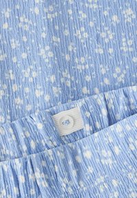 Tissu bleu clair avec un motif moucheté blanc, surface texturée et froissée, et une étiquette blanche avec un trou circulaire au niveau de la ceinture.