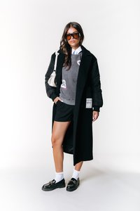 Svart lang jakke over en grå hettegenser med rosa detaljer, svarte shorts og svarte loafers. Har store solbriller og en liten hvit veske.