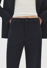 Persoon in zwarte pinstripe broek en blazer met een witte cropped top, handen in de zakken, tegen een effen lichte achtergrond.