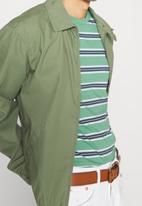 Lekka zielona kurtka z kołnierzem, zestawiona z paskiem zielonym w paski. Pasek wykonany jest z brązowej skóry, a spodnie są białe.