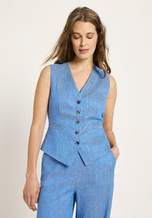 Frau trägt ärmellose hellblaue, geknöpfte Weste und passende Hose, steht mit einer Hand in der Tasche vor schlichtem Hintergrund.