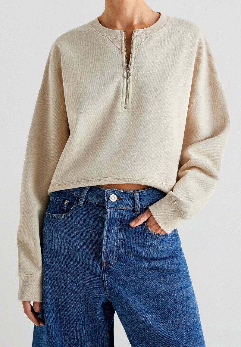 Femme portant un sweat-shirt court beige à manches longues avec fermeture éclair sur le devant et un jean bleu taille haute, main dans la poche.