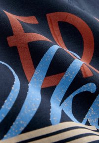 Graafiline T-särk sinise ja oranži tekstiga tumedal kangal. Sile puuvillane tekstuur, millel on kergelt kortsus ilme ja julged tähed.