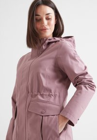 Woman wearing a mauve hooded raincoat with front pockets, one hand in pocket, looking down, against a plain white background.