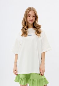 Biały, oversize'owy t-shirt z niebieskim napisem "MIRONS", krótki rękaw, w połączeniu z zieloną spódnicą w falbanki o miękkiej, gładkiej teksturze.