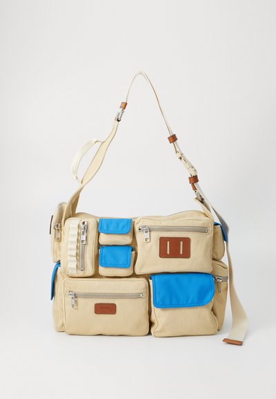 Beige Canvas Tasche mit blauen Akzenten, mehreren Reißverschlussfächern, verstellbarem Schultergurt und braunem Lederetikett.