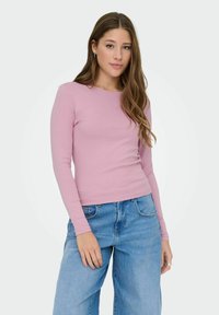 Junge Frau mit langen braunen Haaren, die ein enganliegendes pinkes Langarmoberteil und entspannte blaue Jeans trägt, steht vor einem schlichten Hintergrund.