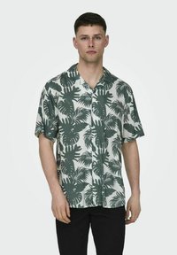 Cămașă cu mânecă scurtă, cu un fond alb și un model de frunze tropicale verzi, având o croială lejeră și guler.