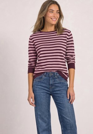 Femme portant un pull rayé violet et rose à manches longues et un jean bleu, souriante et regardant sur le côté sur un fond uni.